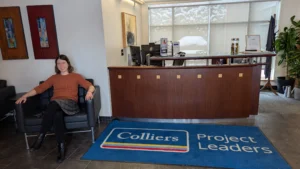 woman with short brown hair wearing an orange sweater sitting in a chair. Beside her is a carpet with text that reads colliers project leaders.